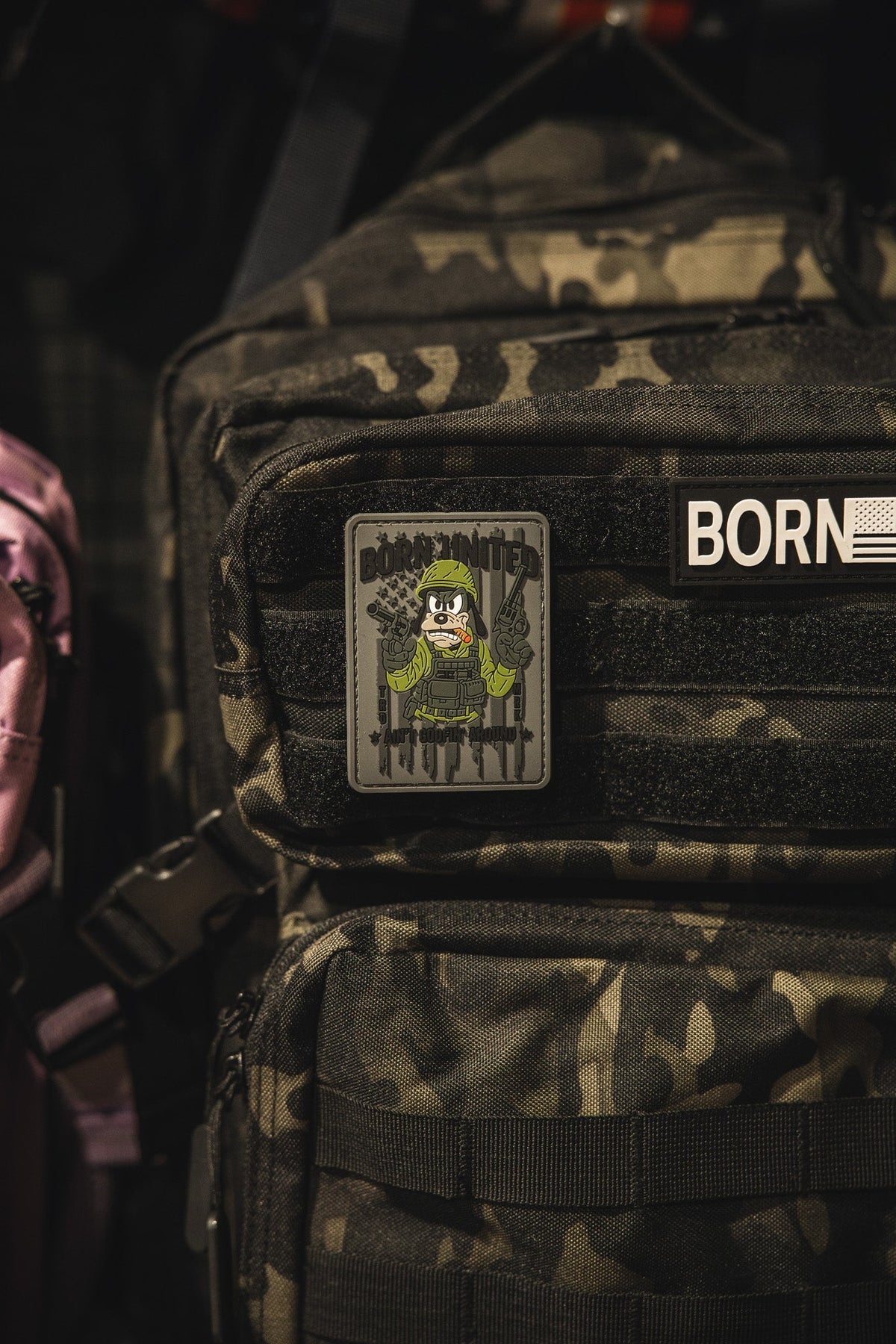 A camouflage backpack features a Born United Private Label Aint Goofin Around PVC patch showing a skeleton in military gear with rifles, attached to the front zip pocket.