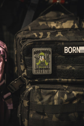 A camouflage backpack features a Born United Private Label Aint Goofin Around PVC patch showing a skeleton in military gear with rifles, attached to the front zip pocket.