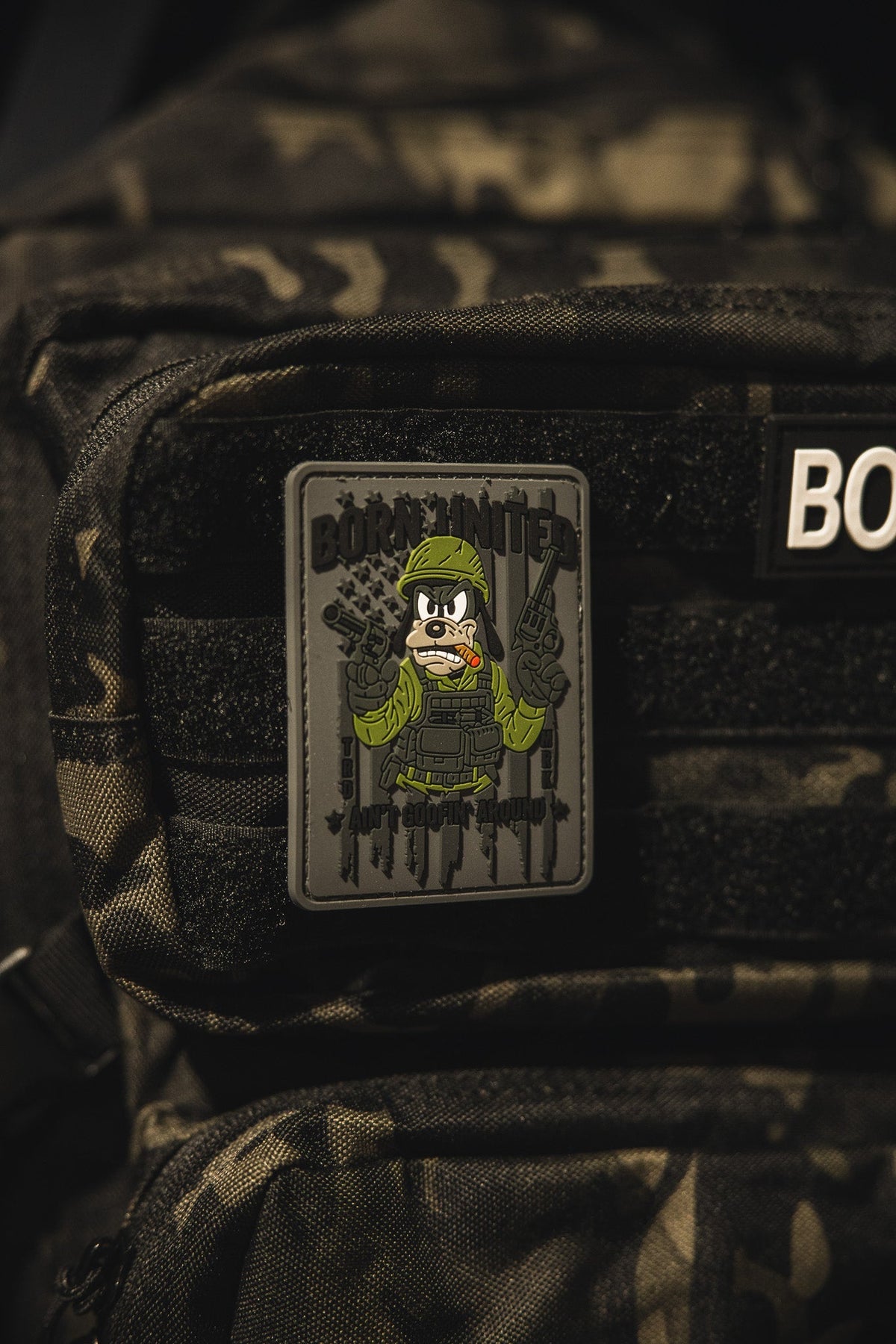 A close-up of a camouflage backpack featuring the Aint Goofin Around PVC Patch from Born United Private Label, which shows a cartoon soldier holding two guns with BORN HUNTED above the character.