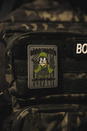 A close-up of a camouflage backpack featuring the Aint Goofin Around PVC Patch from Born United Private Label, which shows a cartoon soldier holding two guns with BORN HUNTED above the character.