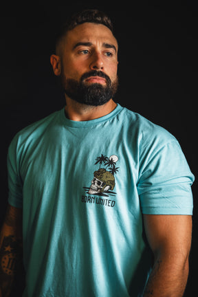 A bearded man wearing The Shallows light blue t-shirt from Born United Private Label, featuring a skull and palm tree graphic, stands against a dark background and looks slightly to the side.