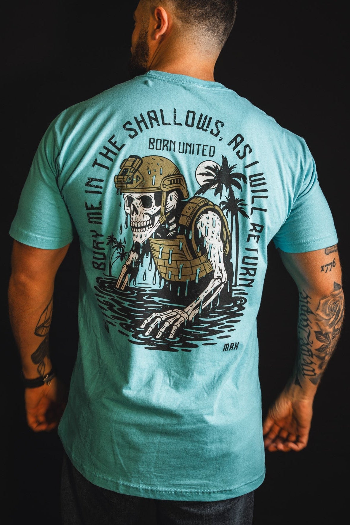 A tattooed man wears "The Shallows" T-shirt by Born United Private Label, featuring a helmeted skeleton rising from water, palm trees, and the text: Bury Me In The Shallows As I Will Return.