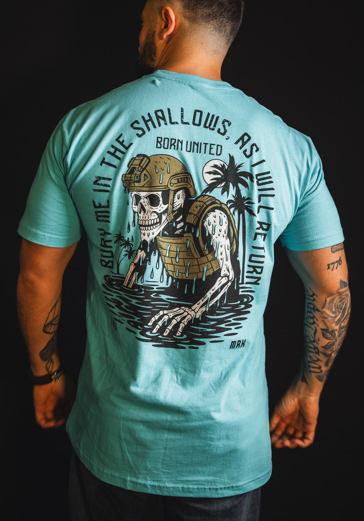 A tattooed man wears "The Shallows" T-shirt by Born United Private Label, featuring a helmeted skeleton rising from water, palm trees, and the text: Bury Me In The Shallows As I Will Return.