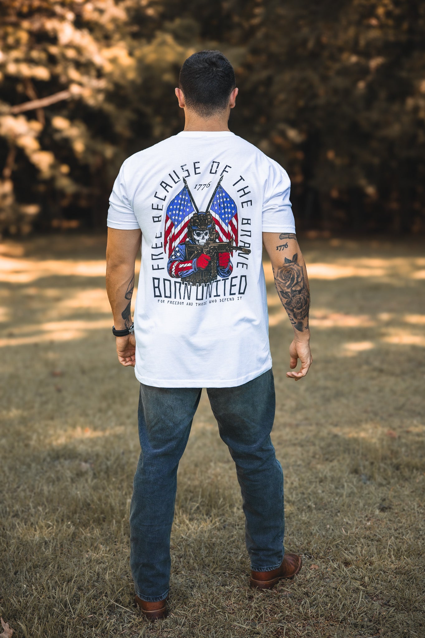 A man stands on grass facing away, wearing a Born United Private Label Freedom Defender white T-shirt featuring an American flag, a helmeted skull, guitars, and "Free Because of the Brave, Born United" on the back. Trees are in the background.