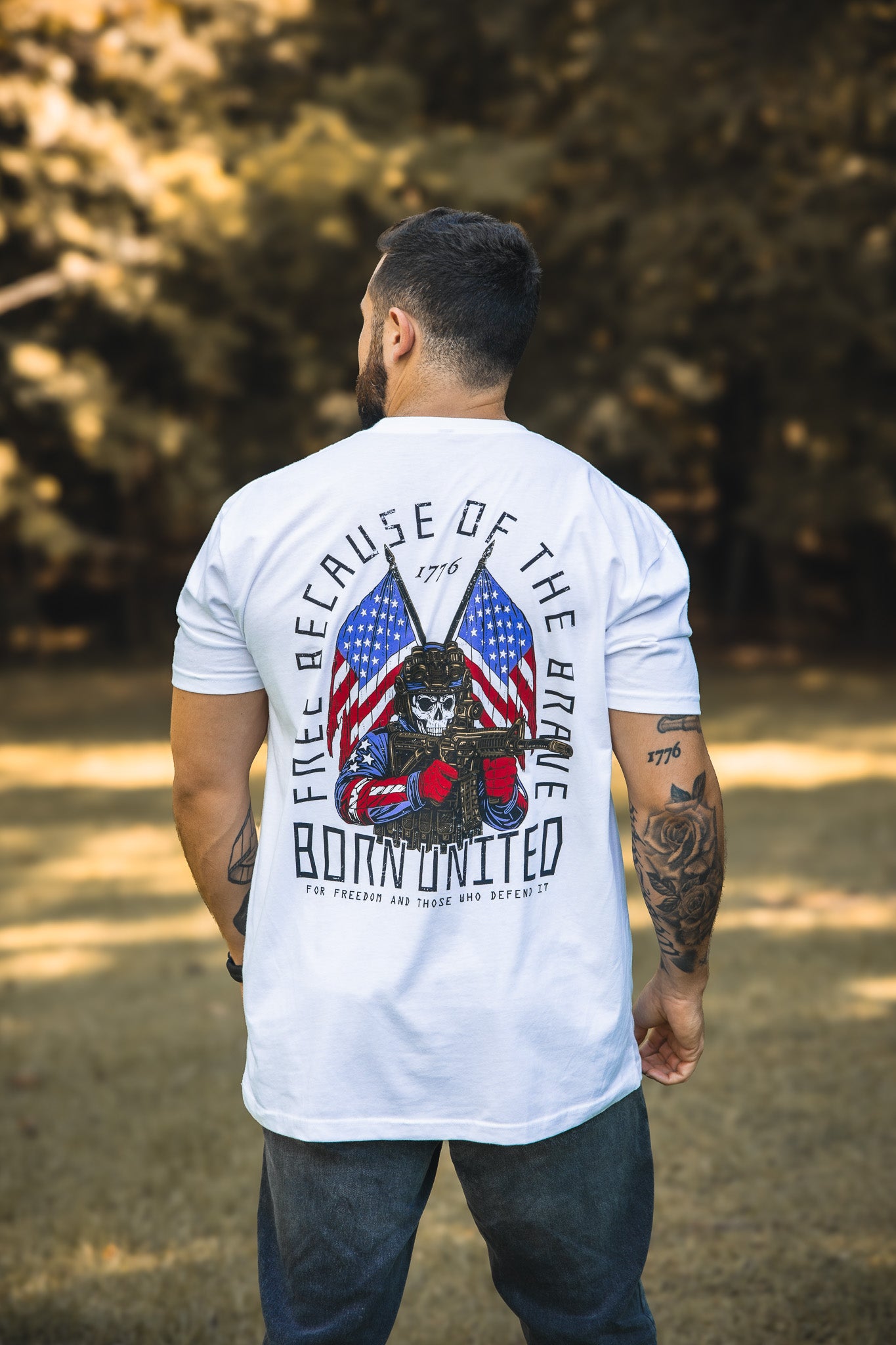 A tattooed man outdoors, back to the camera, wears a white Freedom Defender T-shirt from Born United Private Label featuring a skeleton with boxing gloves, American flags, and the words "Free Because of the Brave – Born United 1776.