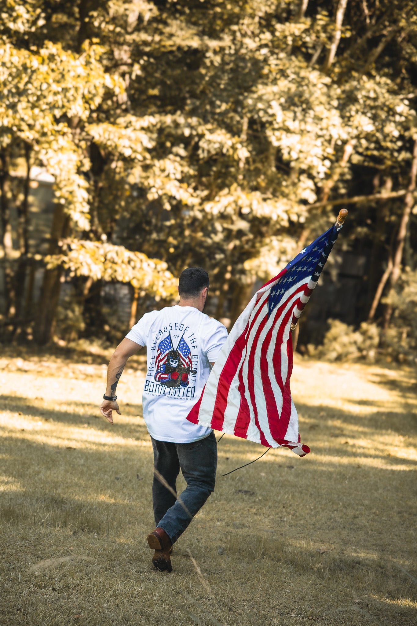 A Freedom Defender from Born United Private Label runs across a grassy field holding an American flag, wearing a white shirt featuring back graphics and text. Lush green trees fill the background.
