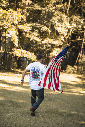 A Freedom Defender from Born United Private Label runs across a grassy field holding an American flag, wearing a white shirt featuring back graphics and text. Lush green trees fill the background.