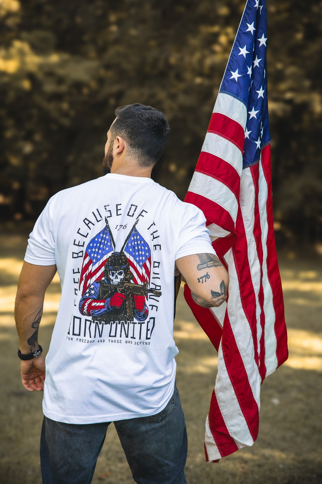 Wearing the Born United Private Label’s Freedom Defender shirt, featuring a skull and American flags with "Free Because of the Brave" and "Born United," a man stands outdoors holding a large American flag, embodying patriotic spirit.