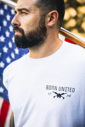 A bearded man in a white "Freedom Defender" t-shirt by Born United Private Label stands outdoors near an American flag, looking thoughtfully into the distance.