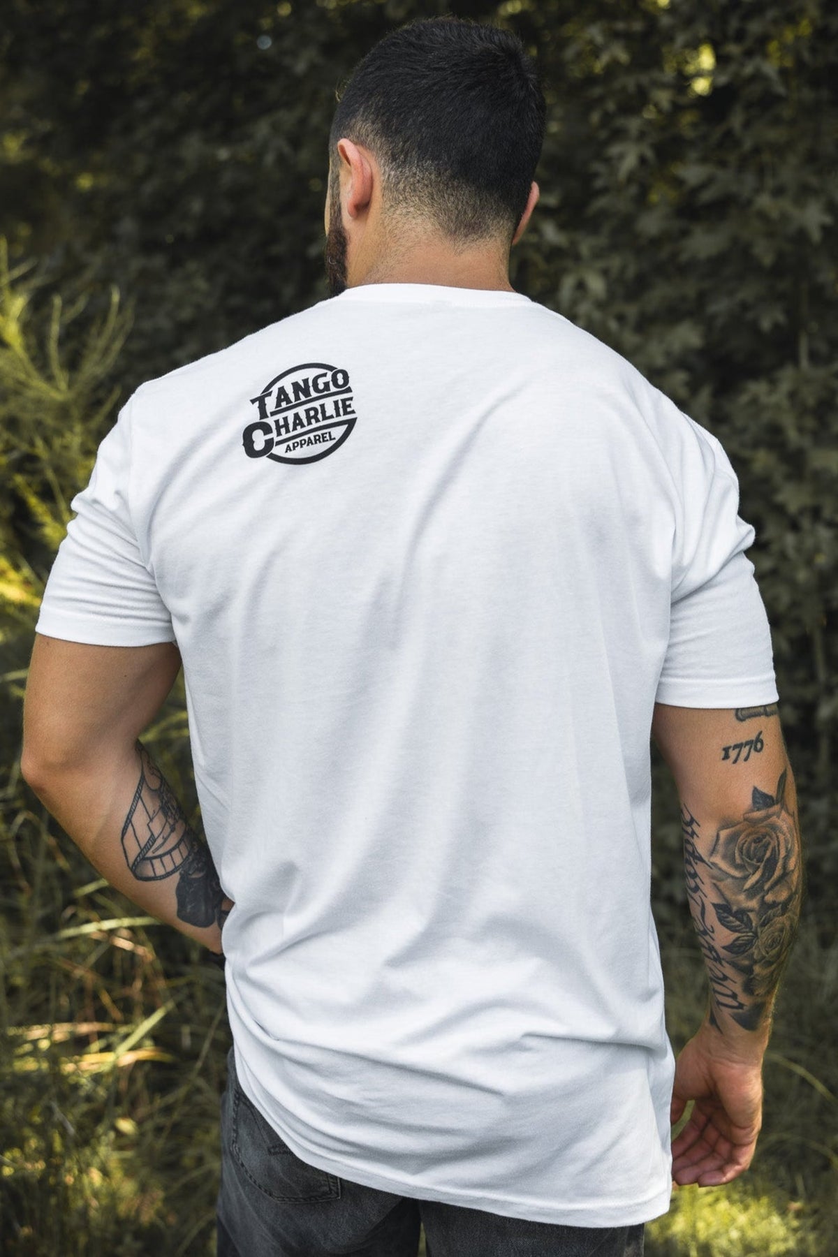 A man stands outdoors with his back to the camera, wearing the Tango Charlie Apparel "Thank You, have a Nice Lift" tee. He shows off his arm tattoos and the soft cotton-poly blend shirt against a backdrop of greenery.