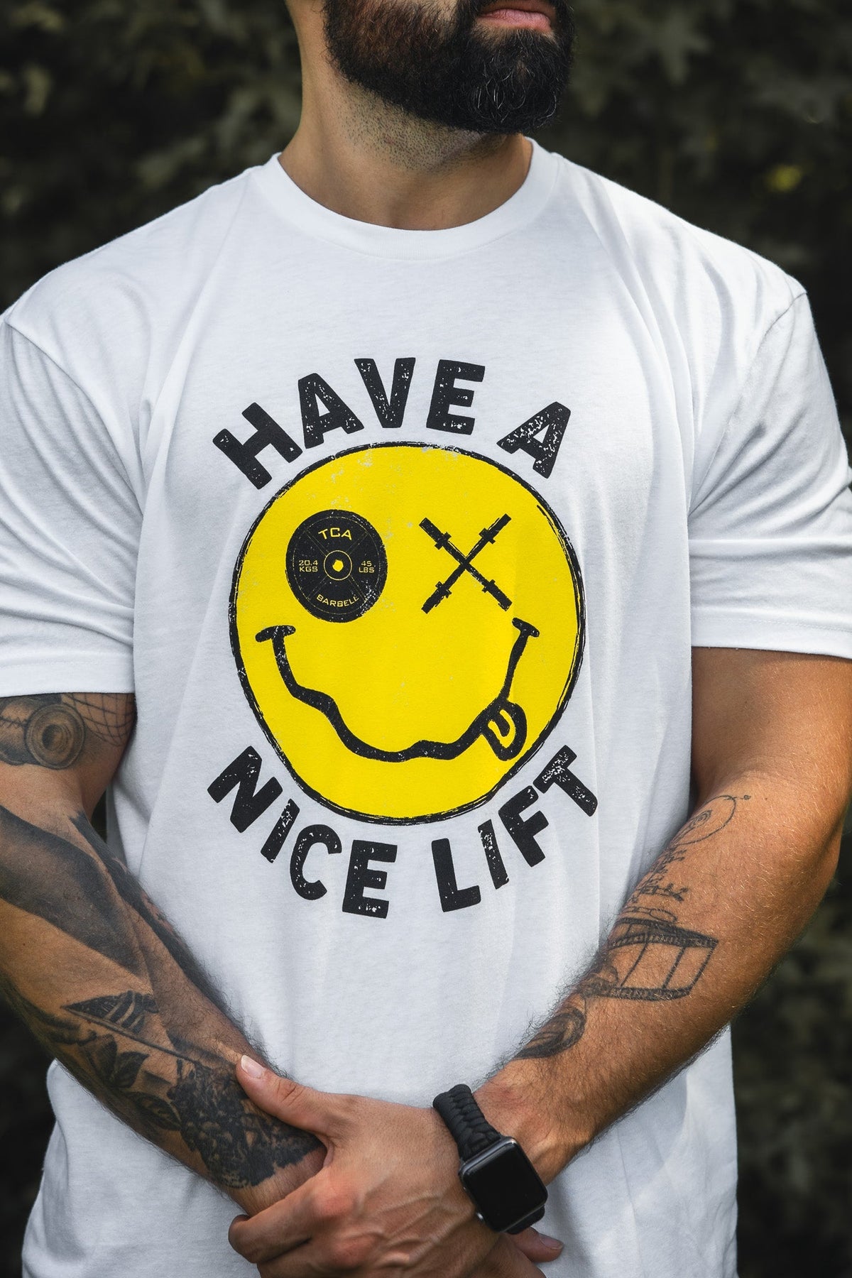 A bearded, tattooed man wears the Tango Charlie Apparel “Thank You, have a Nice Lift" tee, a comfy cotton-poly blend shirt with a yellow smiley face barbell design and the phrase HAVE A NICE LIFT.
