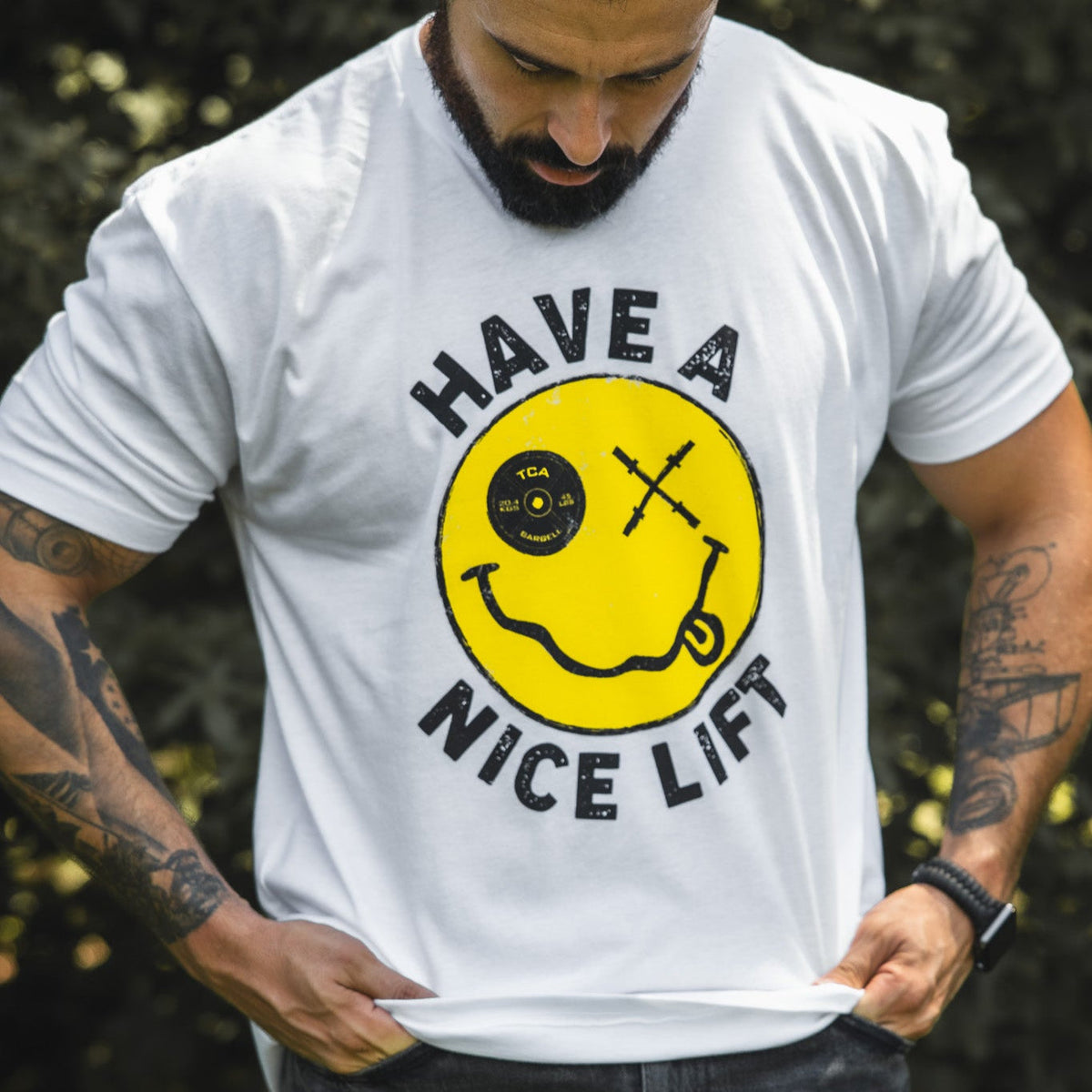 A bearded man looks down at his comfy “Thank You, have a Nice Lift" tee by Tango Charlie Apparel, a white cotton-poly blend with a yellow smiley face featuring a bent barbell mouth, crossed barbell eyes, and “HAVE A NICE LIFT” text.