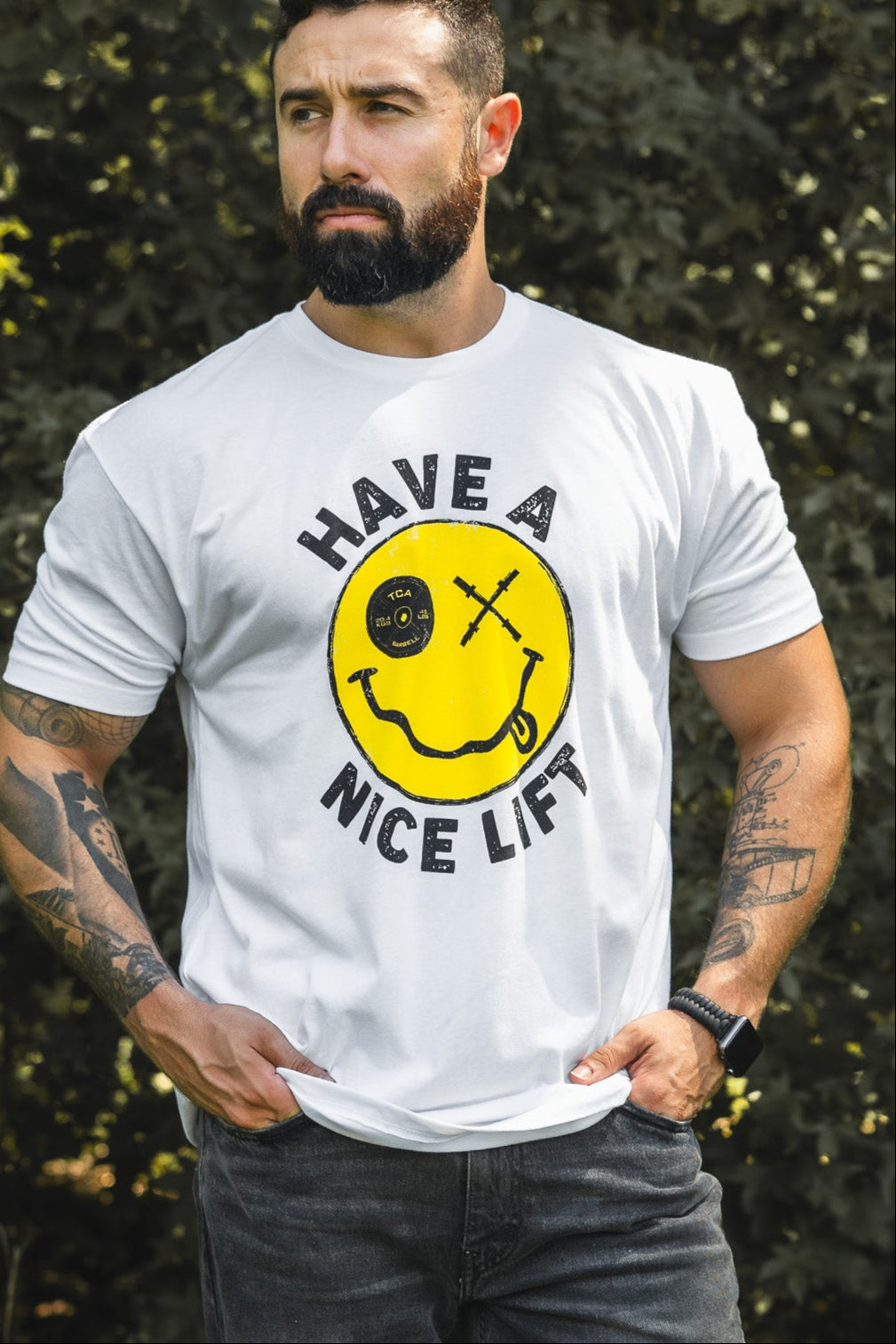 A tattooed, bearded man outdoors wears the Tango Charlie Apparel “Thank You, have a Nice Lift” tee—a comfy white cotton-poly shirt with a yellow smiley face lifting a barbell and the words HAVE A NICE LIFT.