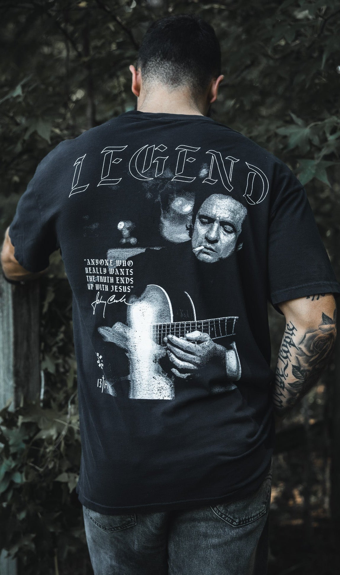 A tattooed man stands outdoors, back turned, wearing the Born United Private Label "Legend - Walk the Line" tee with a grayscale guitar player graphic and the word LEGEND, amid trees and a wooden fence.