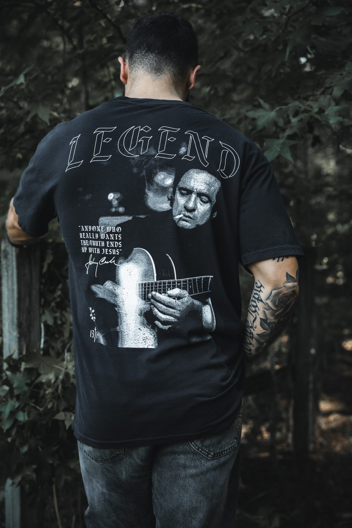A tattooed man stands outdoors, back turned, wearing the Born United Private Label "Legend - Walk the Line" tee with a grayscale guitar player graphic and the word LEGEND, amid trees and a wooden fence.