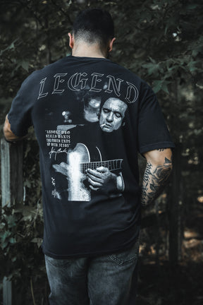 A tattooed man stands outdoors, back turned, wearing the Born United Private Label "Legend - Walk the Line" tee with a grayscale guitar player graphic and the word LEGEND, amid trees and a wooden fence.