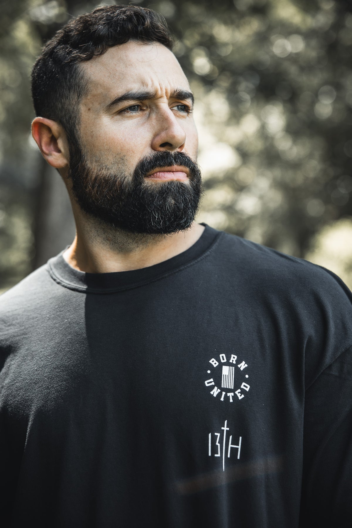 A bearded man with short dark hair stands outdoors wearing the Born United Private Label "Legend - Walk the Line" tee, gazing thoughtfully into the distance with softly blurred trees in the background.