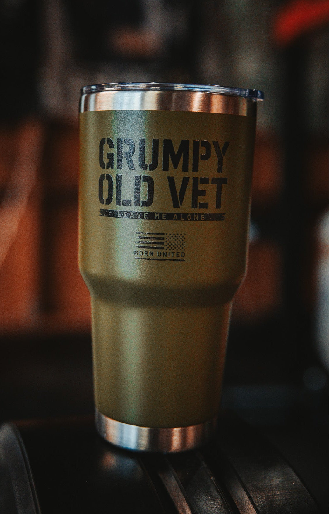 The Grumpy Old Vet Tumbler - 30oz from Born United Private Label, in green with a metal rim, displays bold black text: GRUMPY OLD VET, LEAVE ME ALONE, BORN UNITED against a blurred dark background.