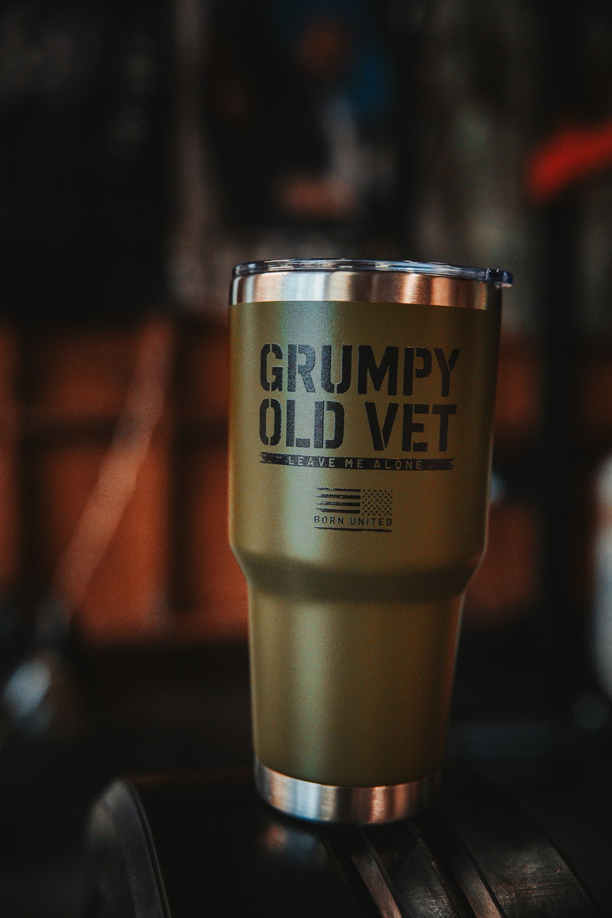 The Grumpy Old Vet Tumbler - 30oz from Born United Private Label, in green with a metal rim, displays bold black text: GRUMPY OLD VET, LEAVE ME ALONE, BORN UNITED against a blurred dark background.
