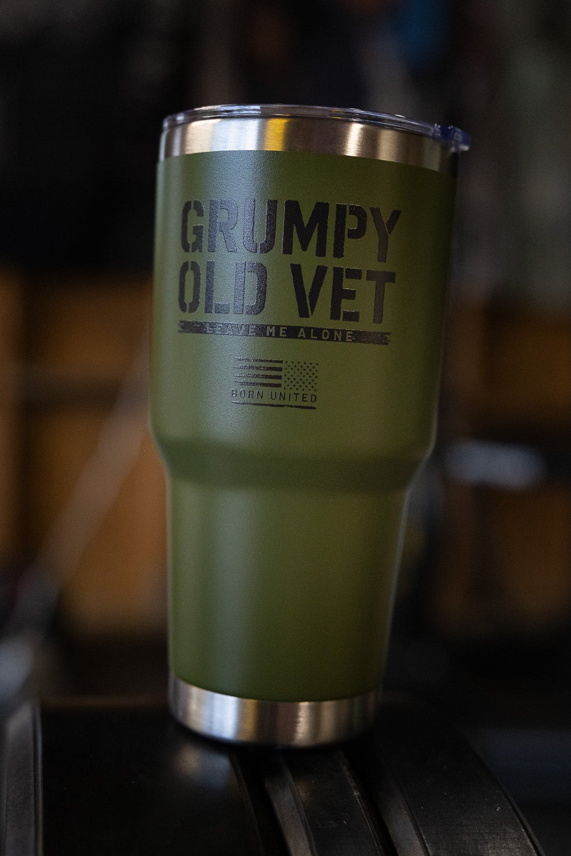 The Born United Private Label Grumpy Old Vet Tumbler - 30oz is green, insulated, and features a metal rim and lid with "GRUMPY OLD VET" on the front, shown on a black surface with a blurred background.