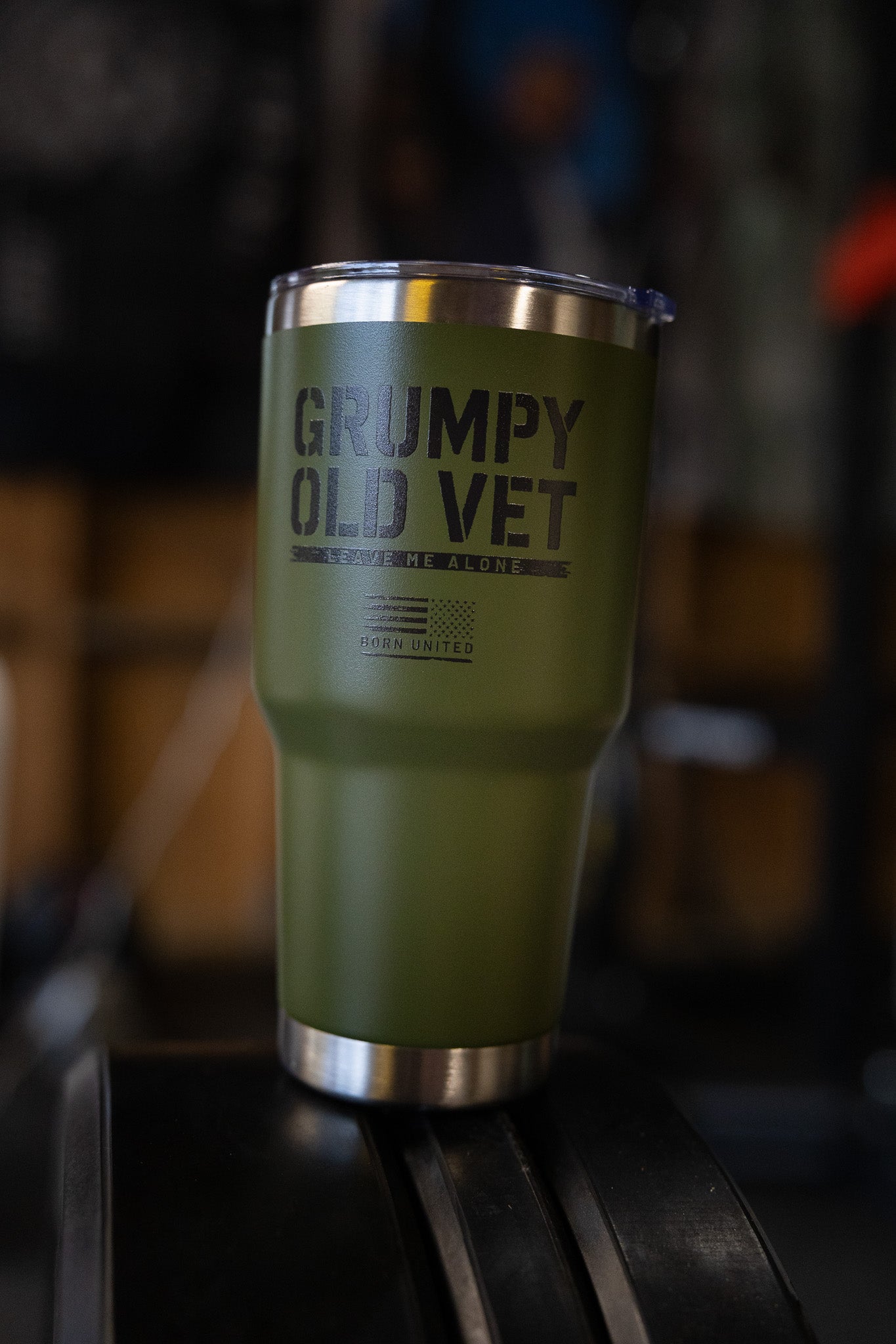 The Born United Private Label Grumpy Old Vet Tumbler - 30oz is green, insulated, and features a metal rim and lid with "GRUMPY OLD VET" on the front, shown on a black surface with a blurred background.