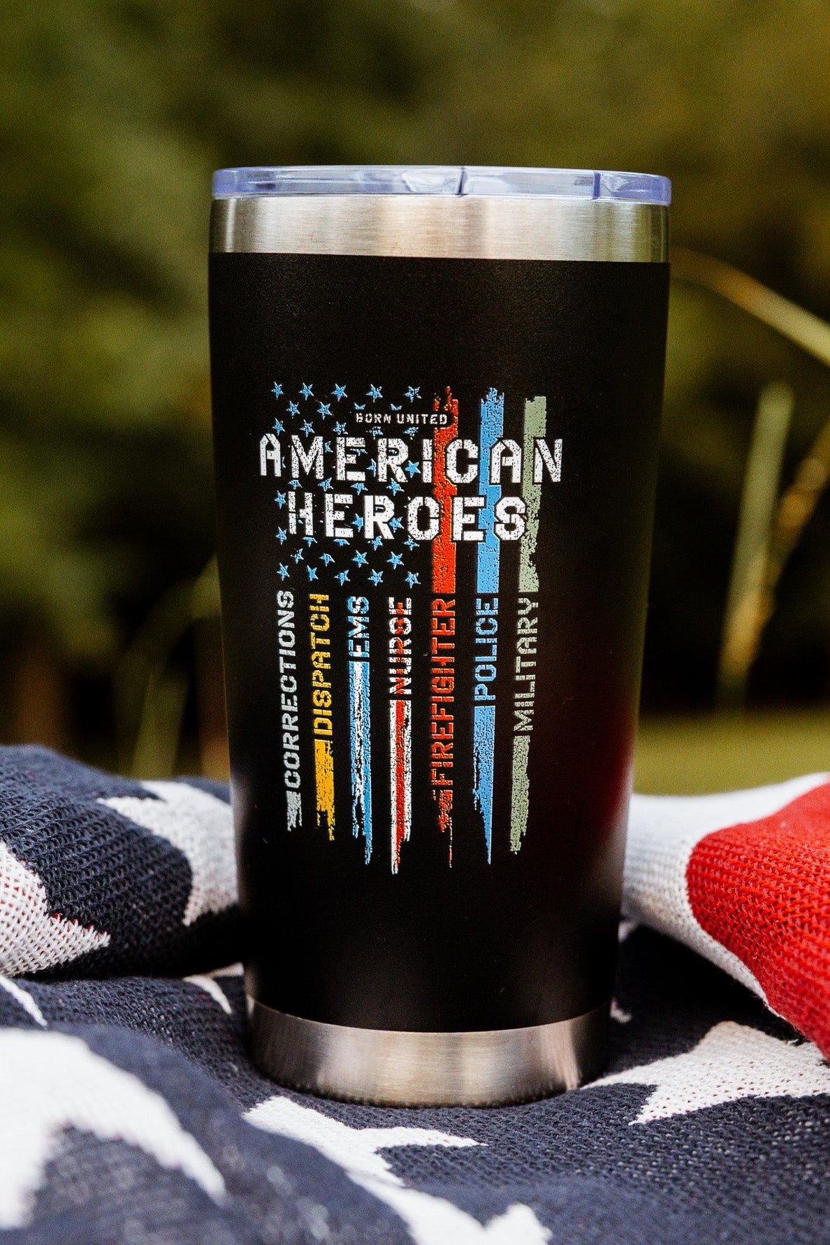 The Born United Private Label American Heroes Tumbler - 20oz features black stainless steel with colorful faded flag stripes, shown on a patriotic blanket outdoors among trees—a striking piece for any patriot.