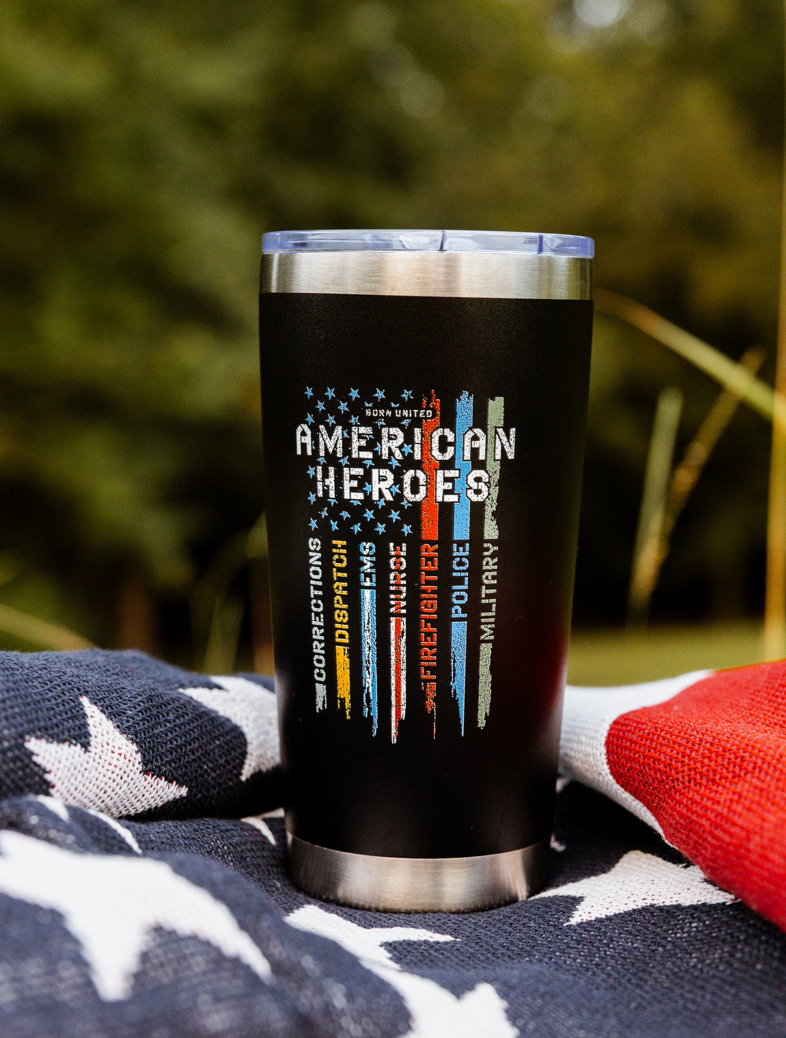 The Born United Private Label American Heroes Tumbler - 20oz features black stainless steel with colorful faded flag stripes, shown on a patriotic blanket outdoors among trees—a striking piece for any patriot.