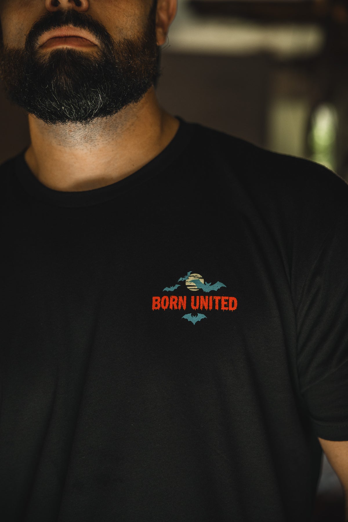 A dark-bearded man wears the Count Tacula limited edition Halloween shirt by Born United Private Label, featuring a small embroidered bat design and "BORN UNITED" in red and orange on the left chest. The image is cropped at his chin.