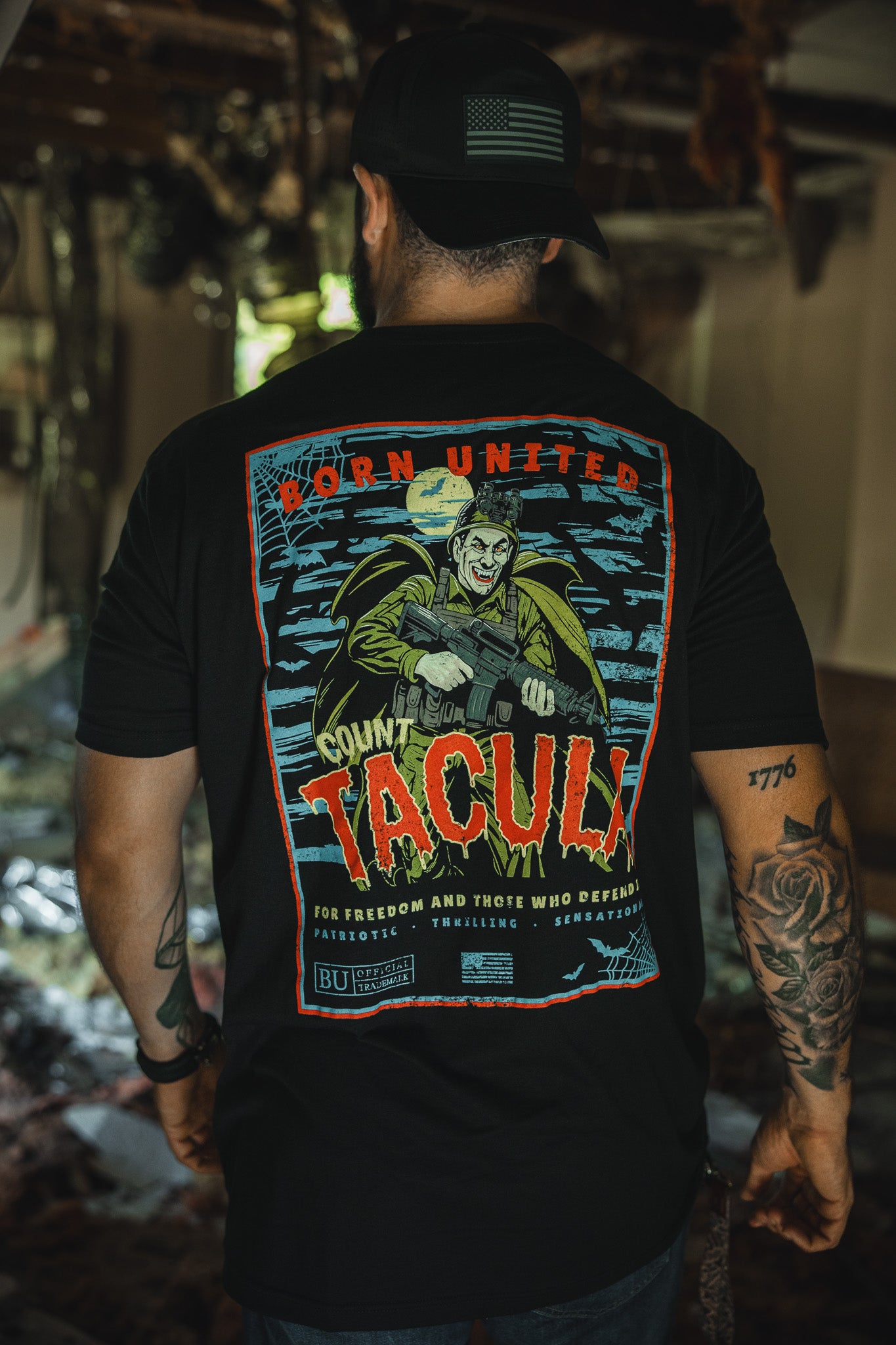 A man faces away indoors, wearing a black cap and the Born United Private Label’s limited edition Count Tacula Halloween shirt with a vibrant Count Tacula holding a rifle and patriotic phrases.