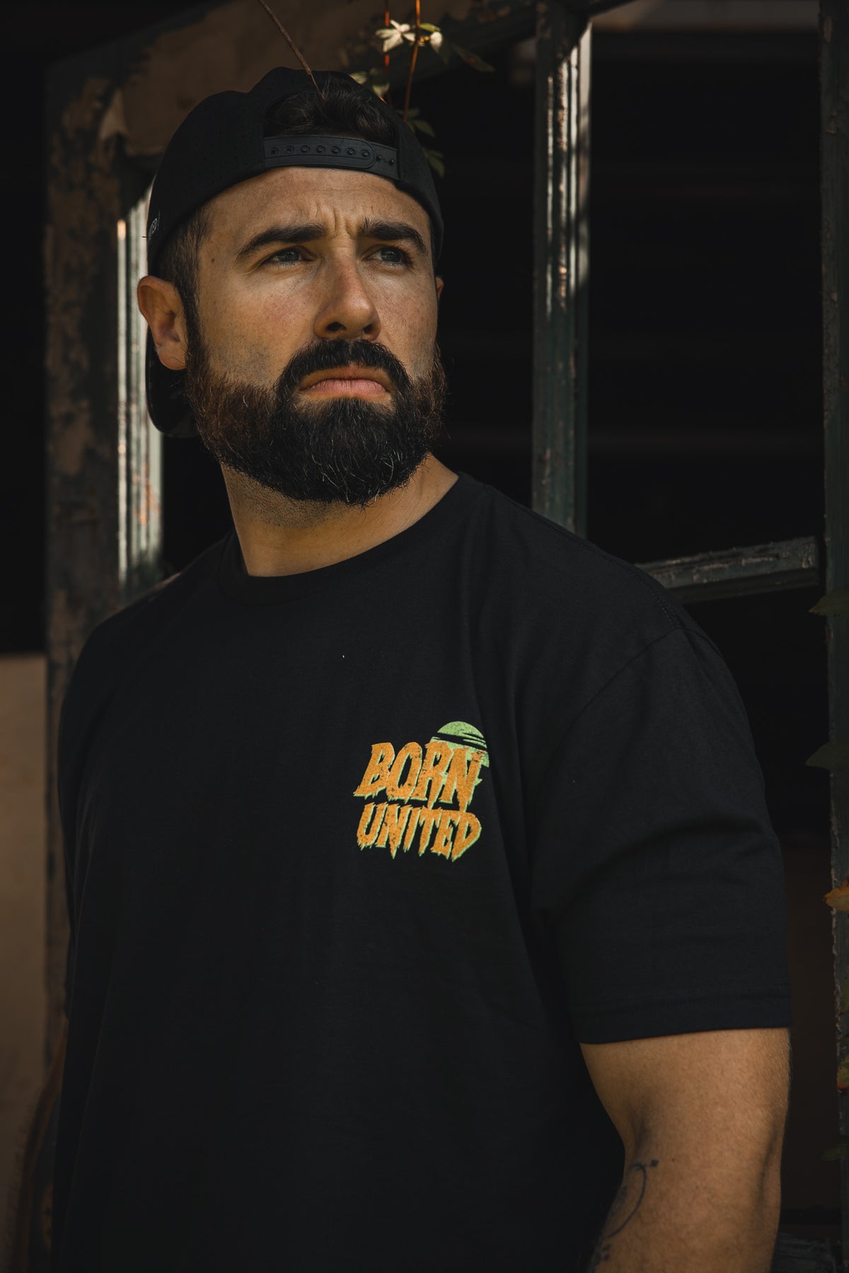 A bearded man in a black backwards cap and a Born United Private Label "Tankenstein" tee stands by a window, looking serious as he gazes into the distance.