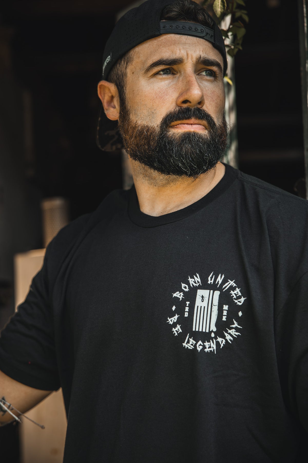 A man with a thick beard and mustache wears the Born United Private Label “Legends of Fear” limited edition black tee with white graphic text and a backward black cap, looking thoughtfully to the side against a blurred background.