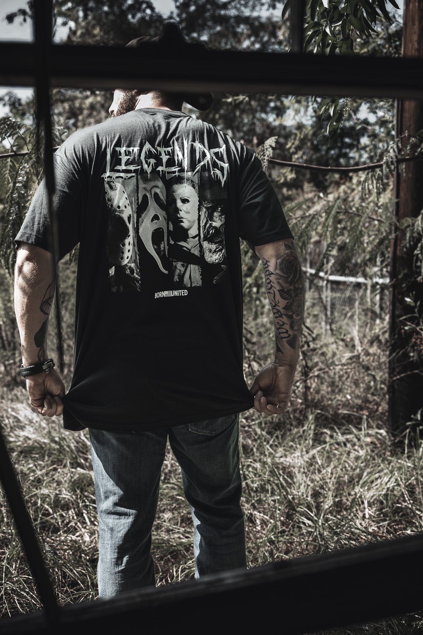 A tattooed man stands in tall grass, wearing Born United Private Label’s “Legends of Fear” tee with iconic horror characters and “LEGENDS” on the back. He faces away as sunlight filters through the trees.