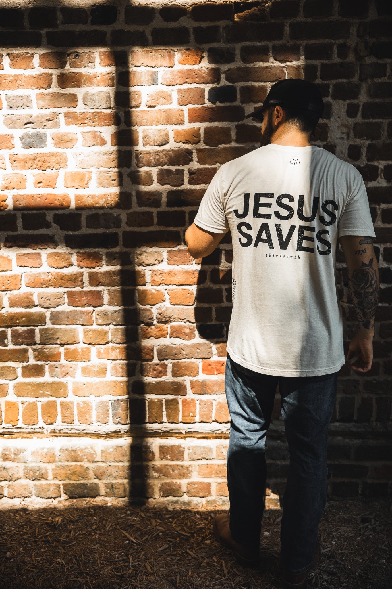 A man in a white "Jesus Saves - White Tee" by 13th faces a brick wall, sunlight casting a cross-shaped shadow beside him—symbolizing John 3:16 and the promise of everlasting life.