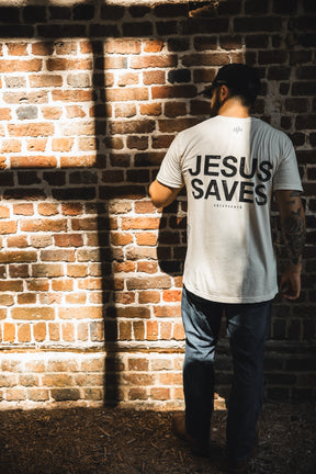 A man in a white "Jesus Saves - White Tee" by 13th faces a brick wall, sunlight casting a cross-shaped shadow beside him—symbolizing John 3:16 and the promise of everlasting life.