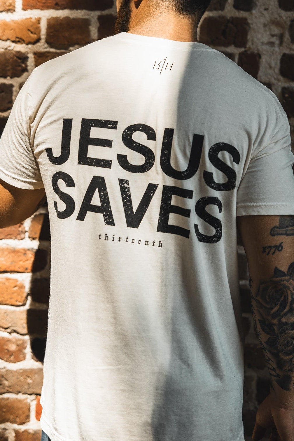 A man faces away from the camera wearing the 13th Jesus Saves - White Tee, its bold message echoing John 3:16 about everlasting life as sunlight casts shadows on the brick wall behind him.