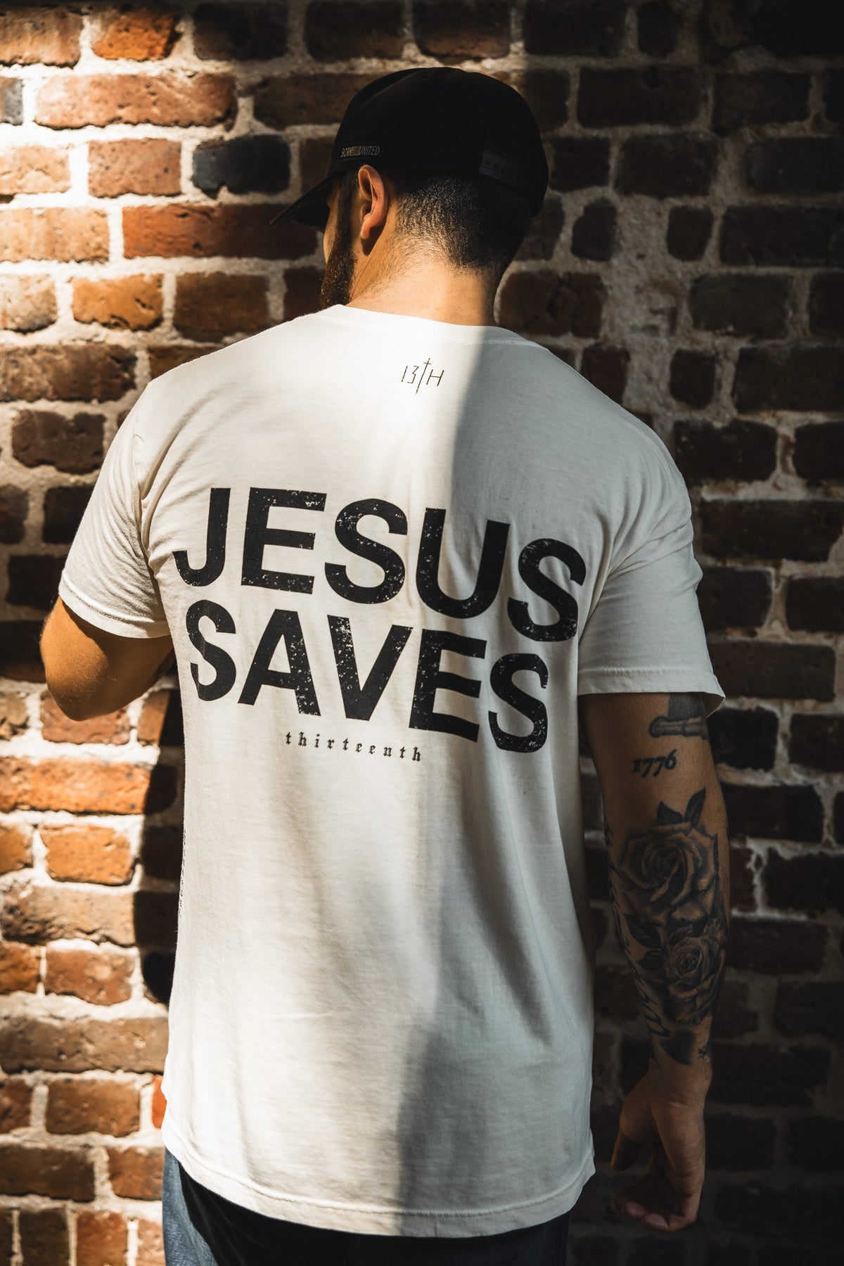 A man faces away from the camera wearing the 13th Jesus Saves - White Tee, its bold message echoing John 3:16 about everlasting life as sunlight casts shadows on the brick wall behind him.