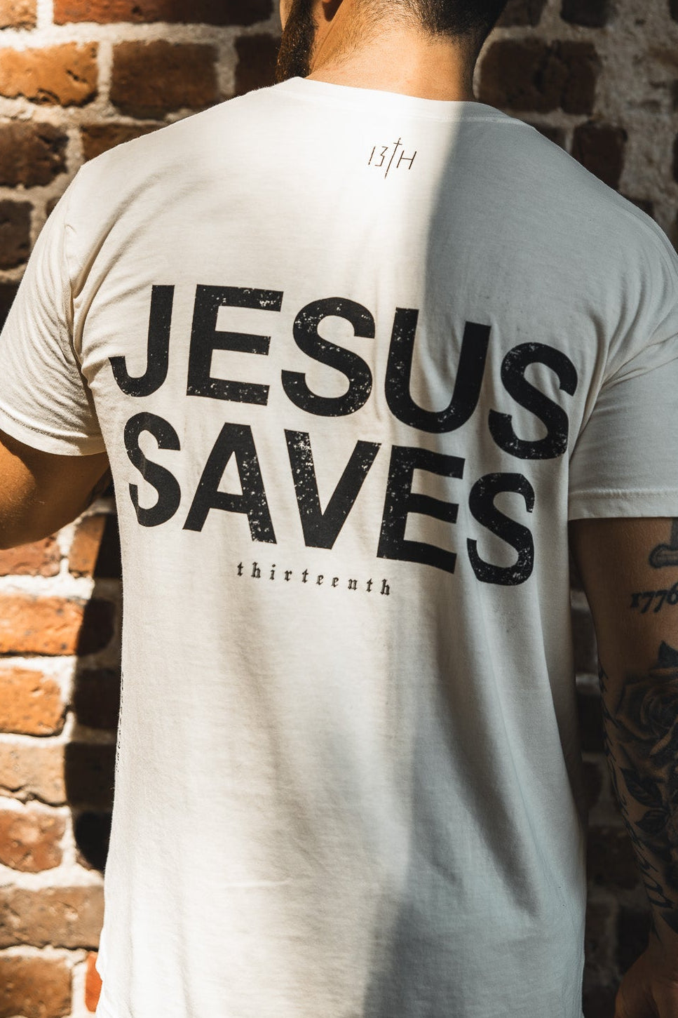 A man faces away from the camera wearing the 13th Jesus Saves - White Tee, its bold message echoing John 3:16 about everlasting life as sunlight casts shadows on the brick wall behind him.