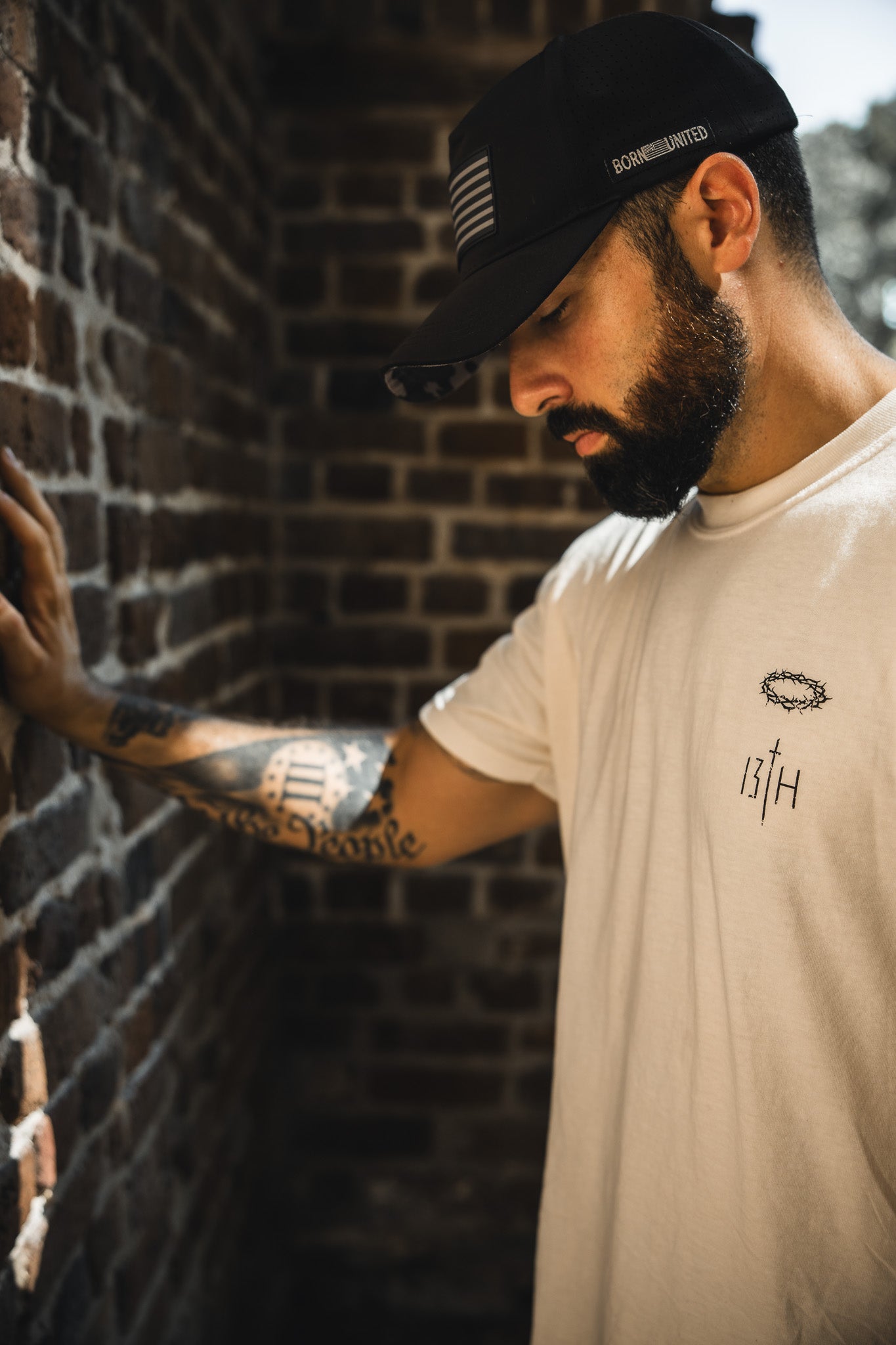 A bearded man in the 13th Jesus Saves White Tee, black cap, and visible Bible verse tattoos leans pensively against a brick wall, sunlight highlighting his face and arm.