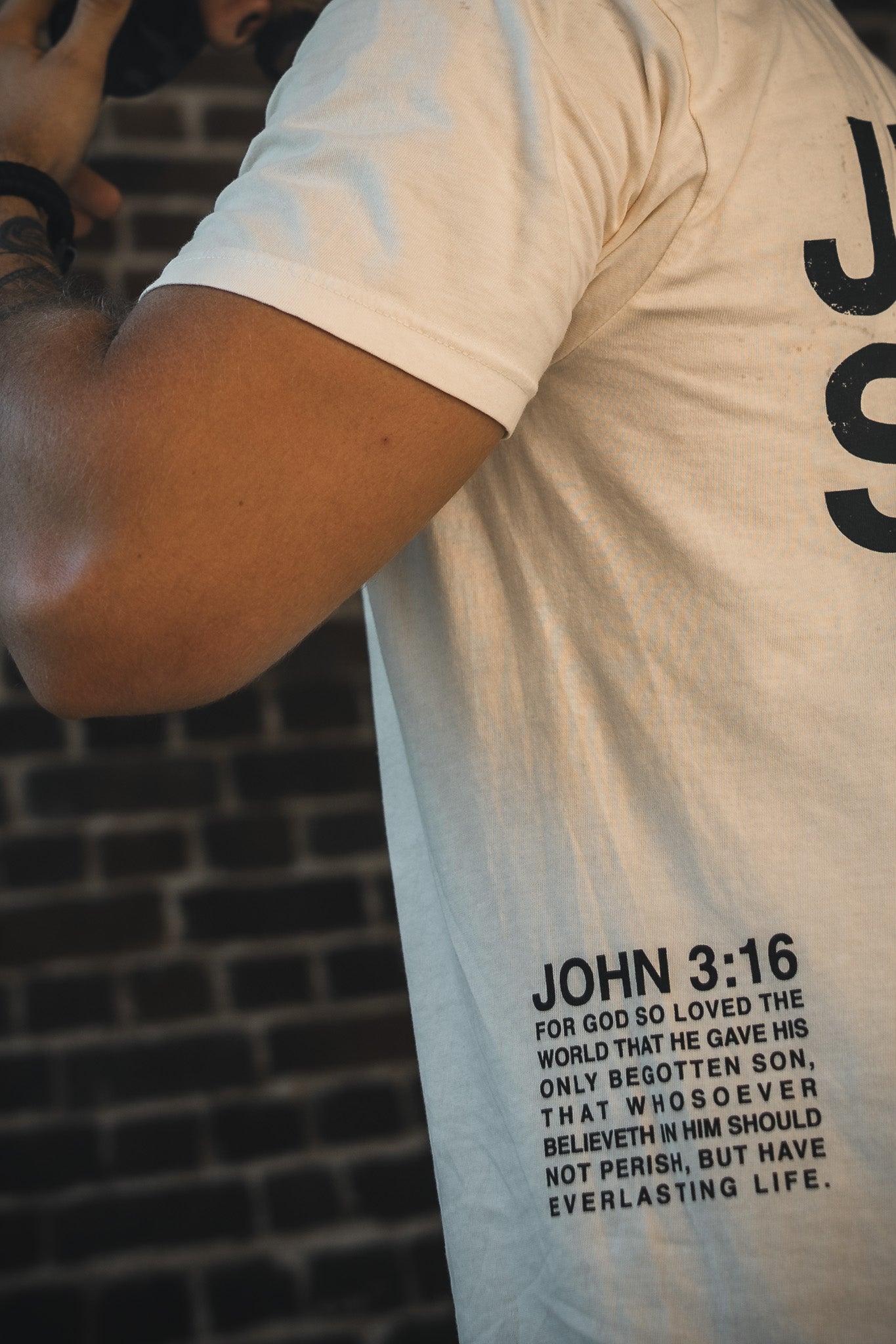 Wearing the 13th Jesus Saves - White Tee, a person stands before a brick wall, with John 3:16 and its message of everlasting life displayed on their back; only part of their upper body and arm are visible.
