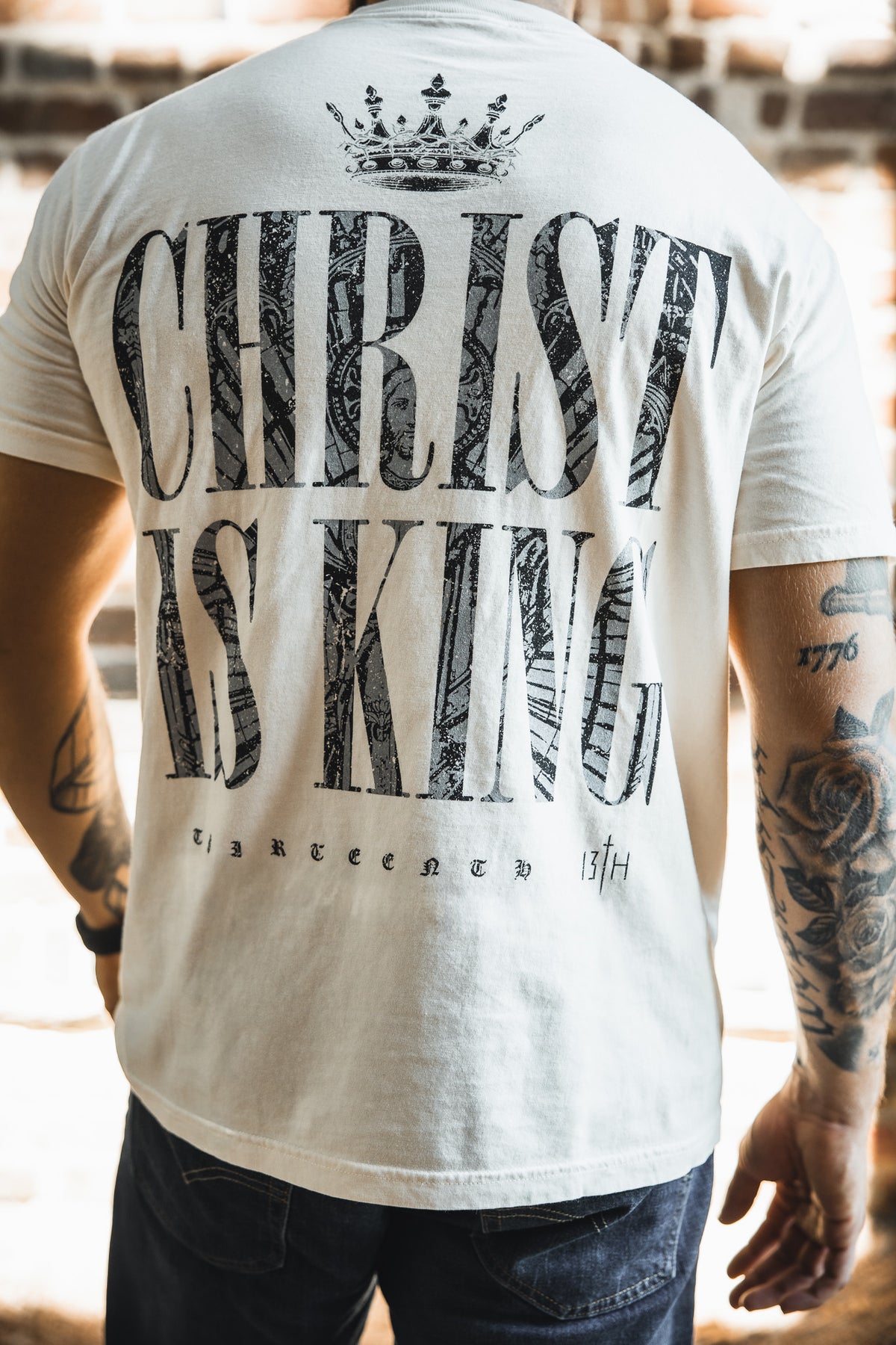 A person wearing the Christ is King Premium Tee - Ivory by 13th, featuring bold "CHRIST IS KING" text, a crown graphic above, and "thirteen 13th" below. Visible arm tattoos complement this standout Christian shirt.