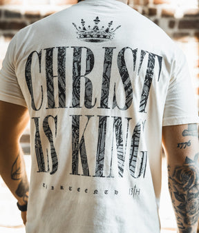 A person wearing the Christ is King Premium Tee - Ivory by 13th, featuring bold "CHRIST IS KING" text, a crown graphic above, and "thirteen 13th" below. Visible arm tattoos complement this standout Christian shirt.