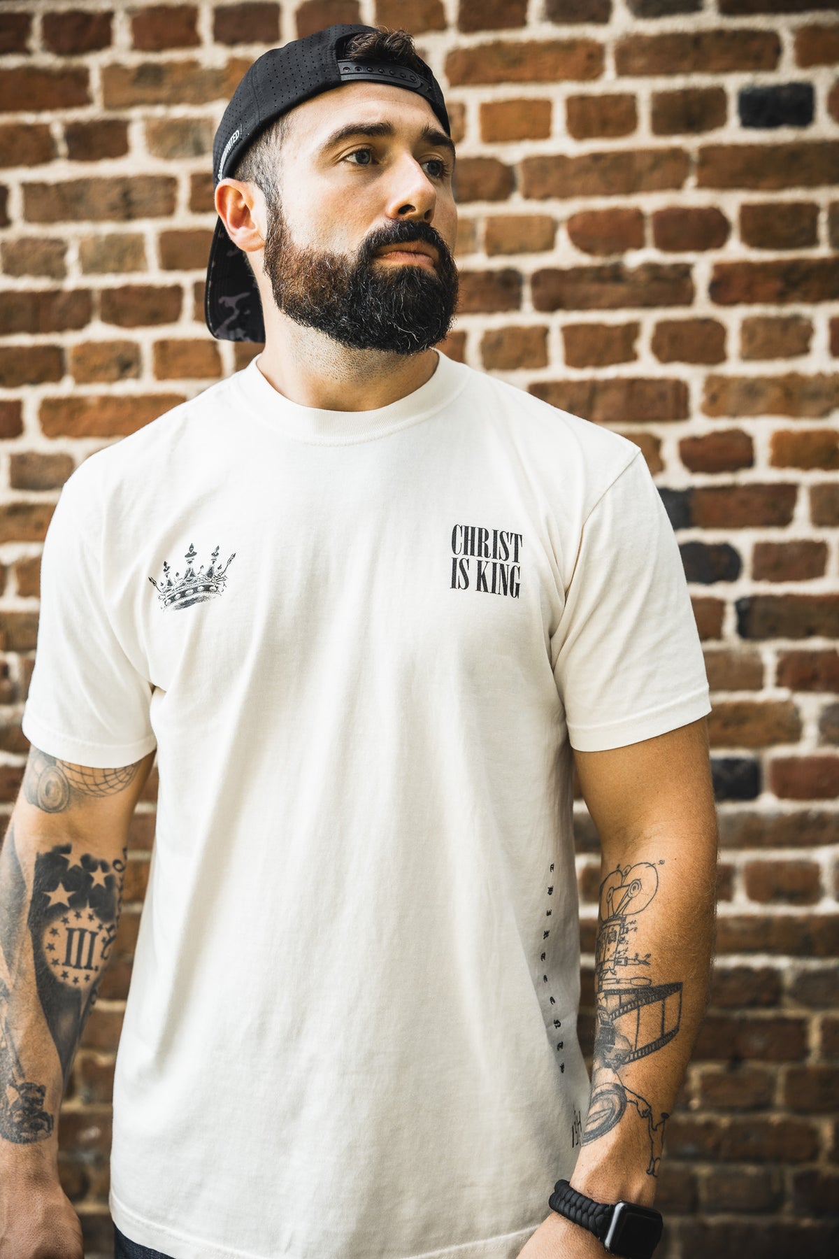 A bearded man in a backwards cap wears the 13th Christ is King Premium Tee - Ivory, standing before a brick wall with tattooed arms and a neutral expression, looking off to the side.