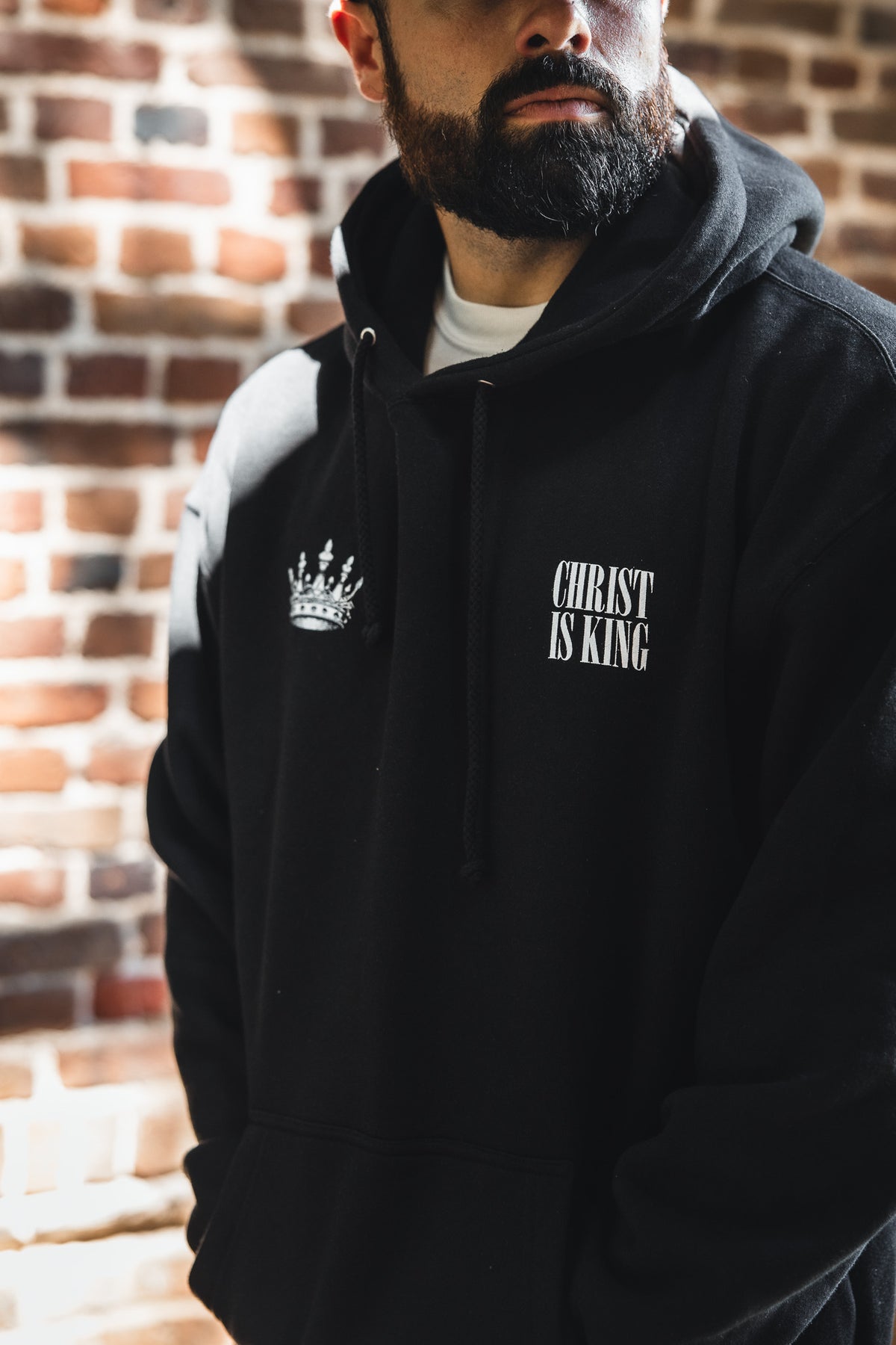A bearded man in front of a brick wall wears the 13th brand’s Christ is King Premium Hoodie, which is black and features “CHRIST IS KING” with a crown graphic.