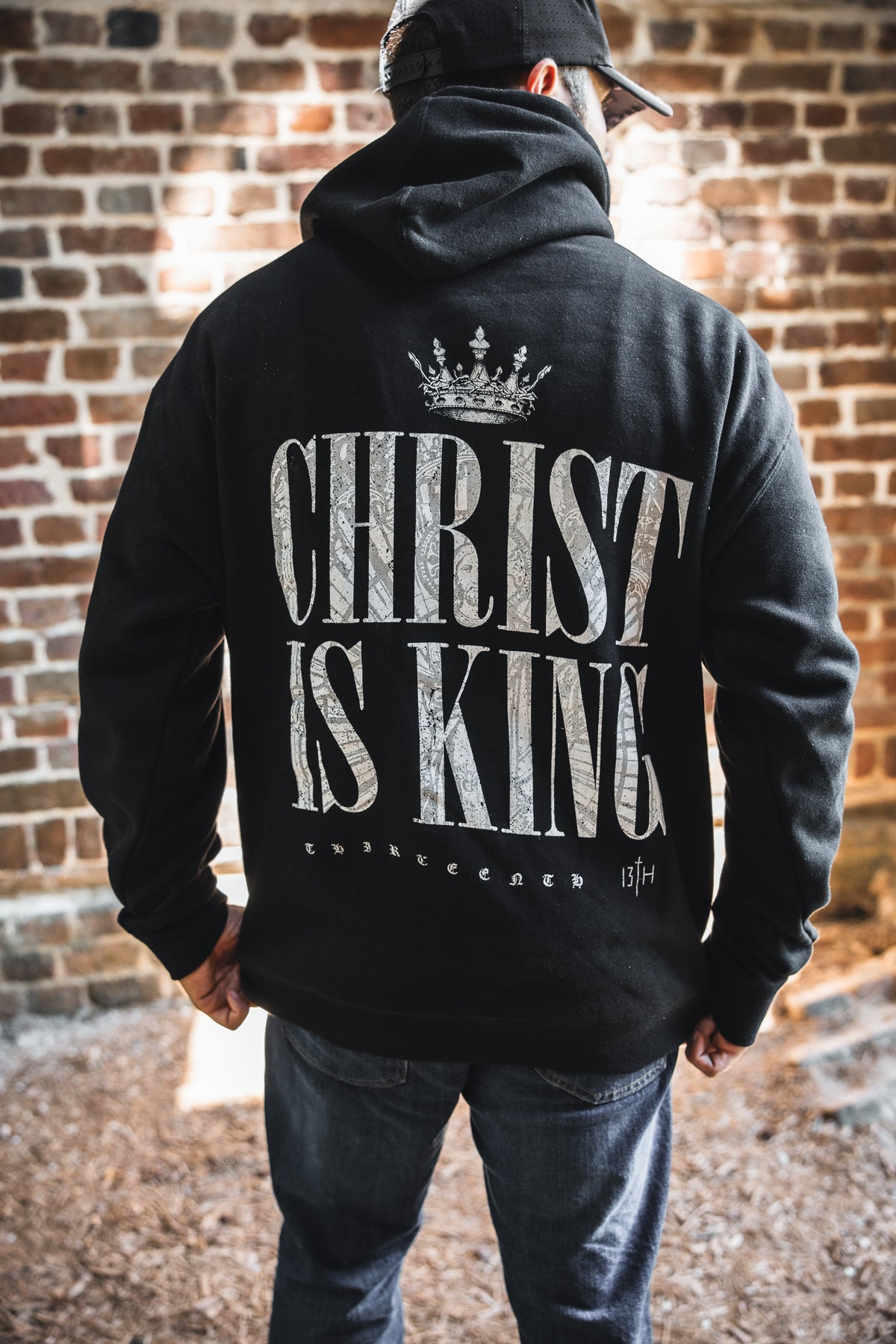 Wearing the 13th Christ is King Premium Hoodie—a black hoodie with bold "CHRIST IS KING" text and a crown—a person stands outside facing a brick wall, turning their back to showcase the faith-inspired design.