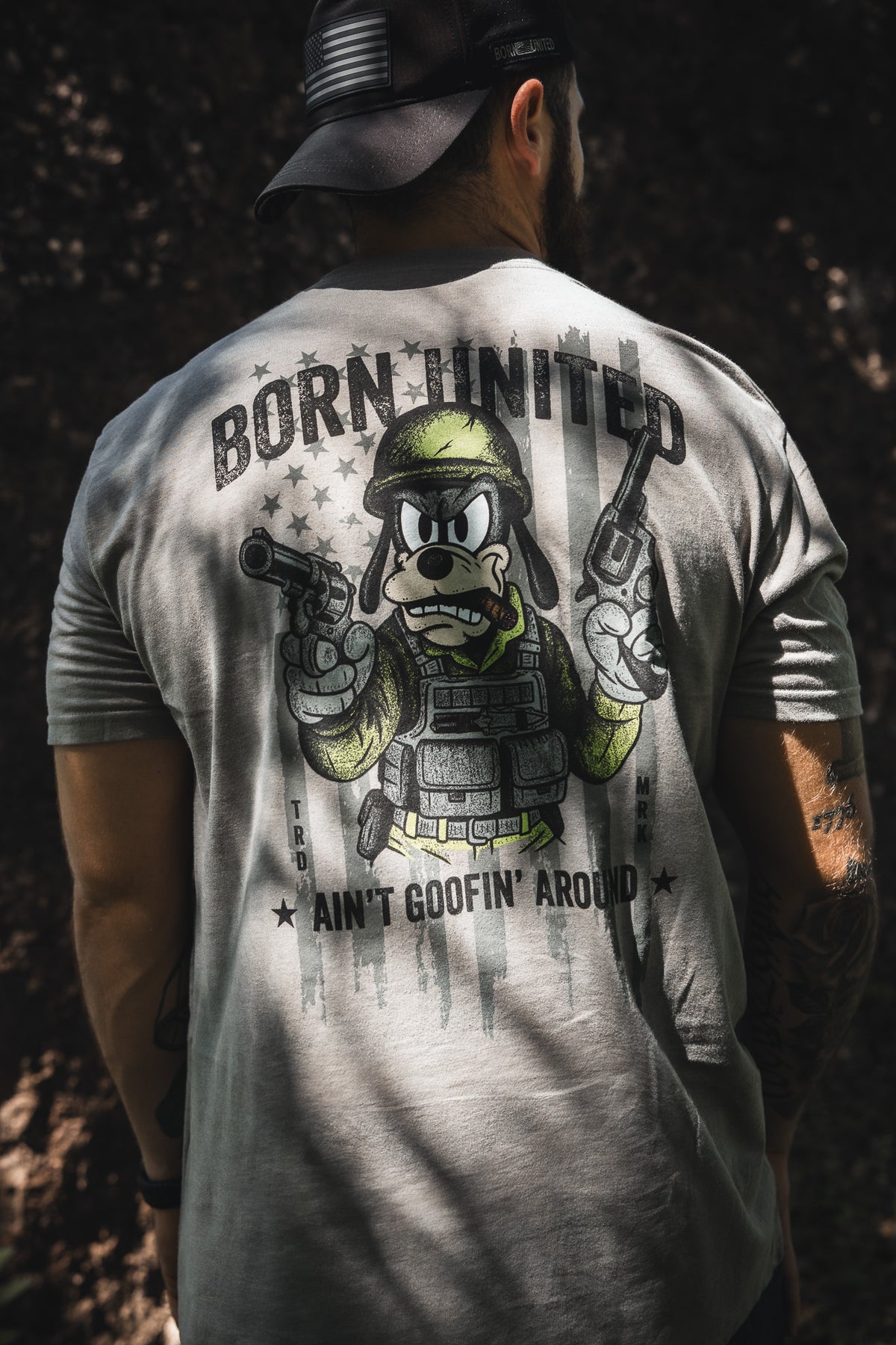 A man wearing a cap faces away, showing the "Ain't Goofin' Around" T-shirt by Born United Private Label, with a cartoon dog in military gear holding pistols. “BORN UNITED” is above and “AIN'T GOOFIN' AROUND” is boldly below the image.