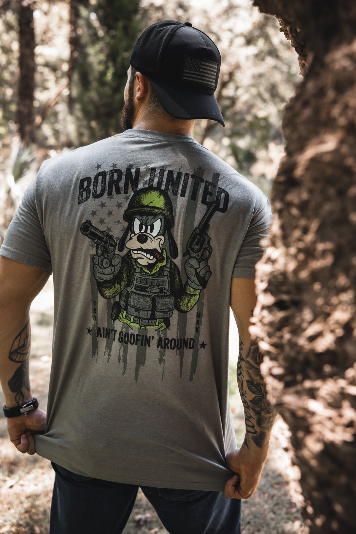 A man wearing a gray "Ain't Goofin' Around" t-shirt by Born United Private Label, featuring a cartoon soldier and pistols, stands outdoors among trees, viewed from behind while sporting a black cap.