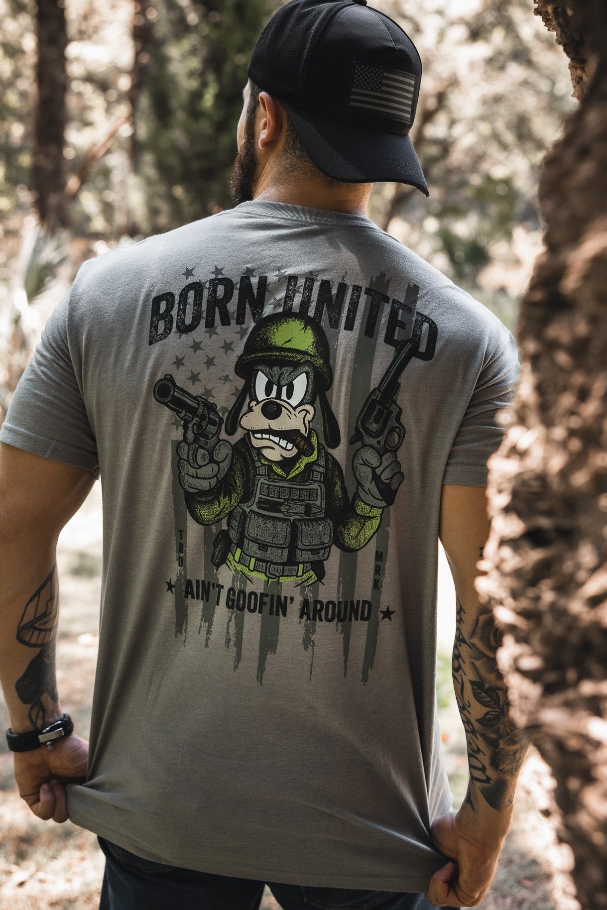 A man wearing a gray "Ain't Goofin' Around" t-shirt by Born United Private Label, featuring a cartoon soldier and pistols, stands outdoors among trees, viewed from behind while sporting a black cap.