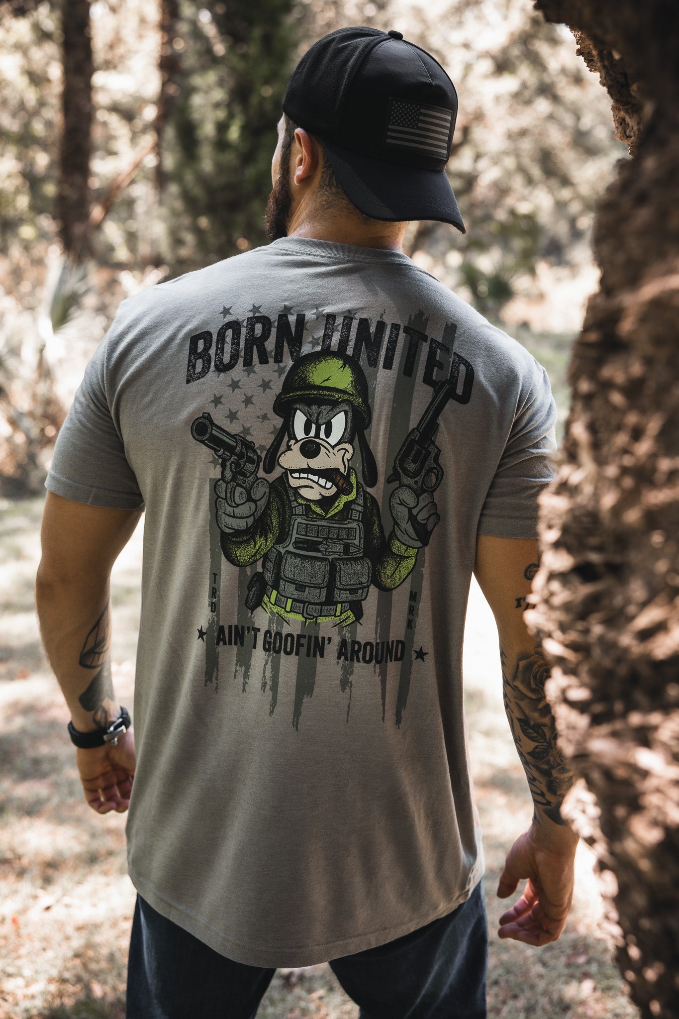 A man in a black cap and gray Born United Private Label “Ain’t Goofin’ Around” T-shirt stands outdoors; the shirt’s back displays a cartoon soldier with guns, bold text, and a faded American flag.