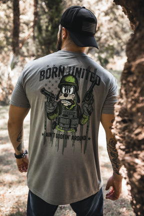 A man in a black cap and gray Born United Private Label “Ain’t Goofin’ Around” T-shirt stands outdoors; the shirt’s back displays a cartoon soldier with guns, bold text, and a faded American flag.