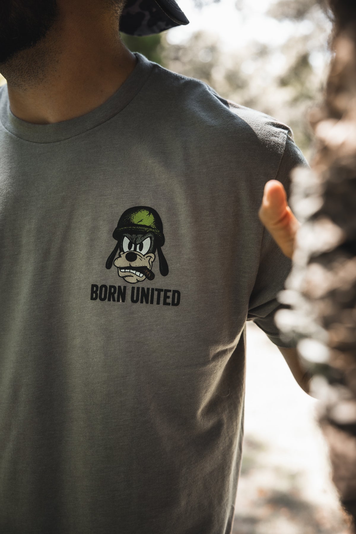 A person wears the "Ain't Goofin' Around" gray T-shirt from Born United Private Label, featuring a cartoon dog in a green helmet and "BORN UNITED" text on the chest, outdoors in sunlight.